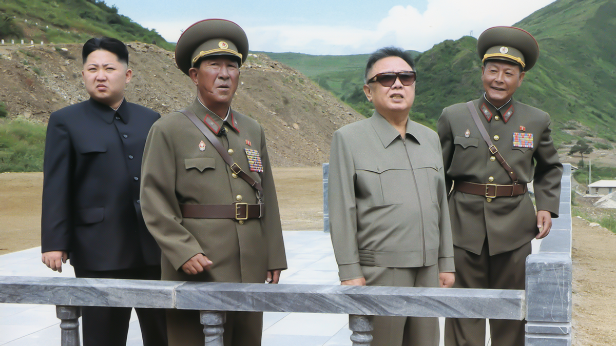 Four Asian people stand next to each other on a white platform. A person wearing a military outflit is next to one person wearing a dark blue suit, while the person standing next to the other military person is wearing a gray outfit. 