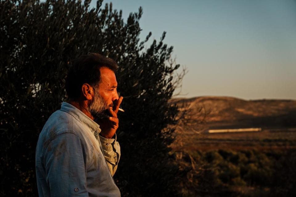 A man pictured from the chest up, standing beside a tree and smoking a cigarette as he looks out at surrounding land