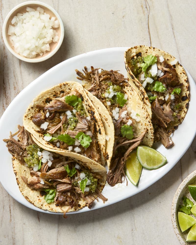 overhead shot of pork carnitas tacos on a white plate, with onion, lime and salsa on the side.