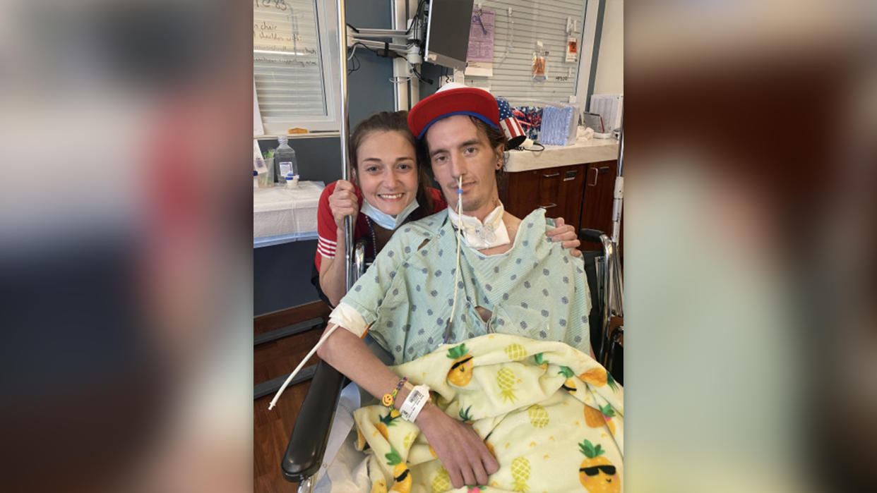  A young man seated and wearing a hospital gown and hat pictured next to his partner in a hospital room. 