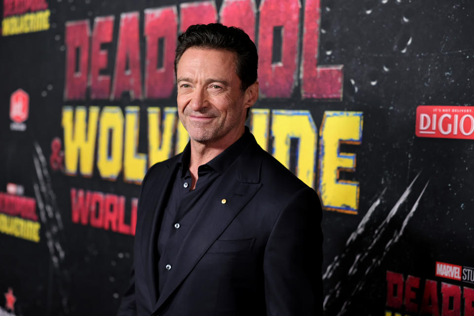 Hugh Jackman stands in front of a "Deadpool & Wolverine" event backdrop, wearing a dark suit