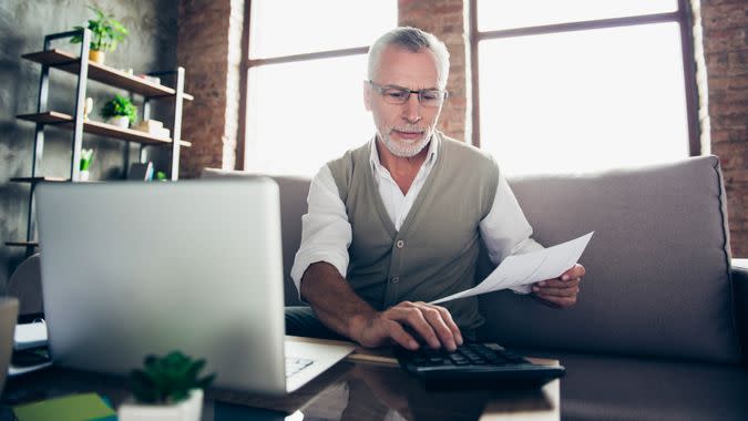 Qualified experienced intelligent confident concentrated old bearded man checking his calculations at home using calculator and netbook.