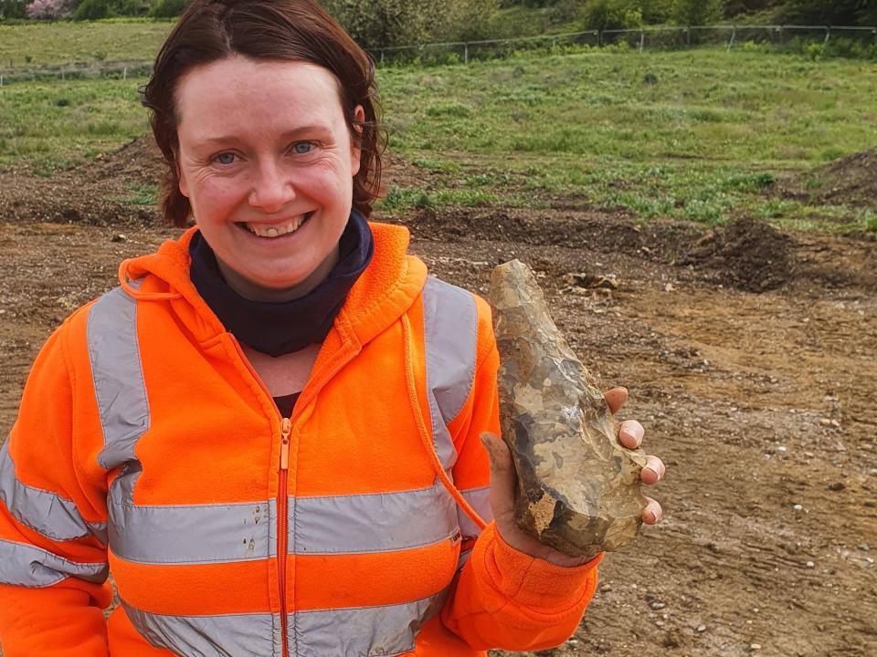 A woman in an orange safety jacket smiles as she holds up a large handaxe to the camera.