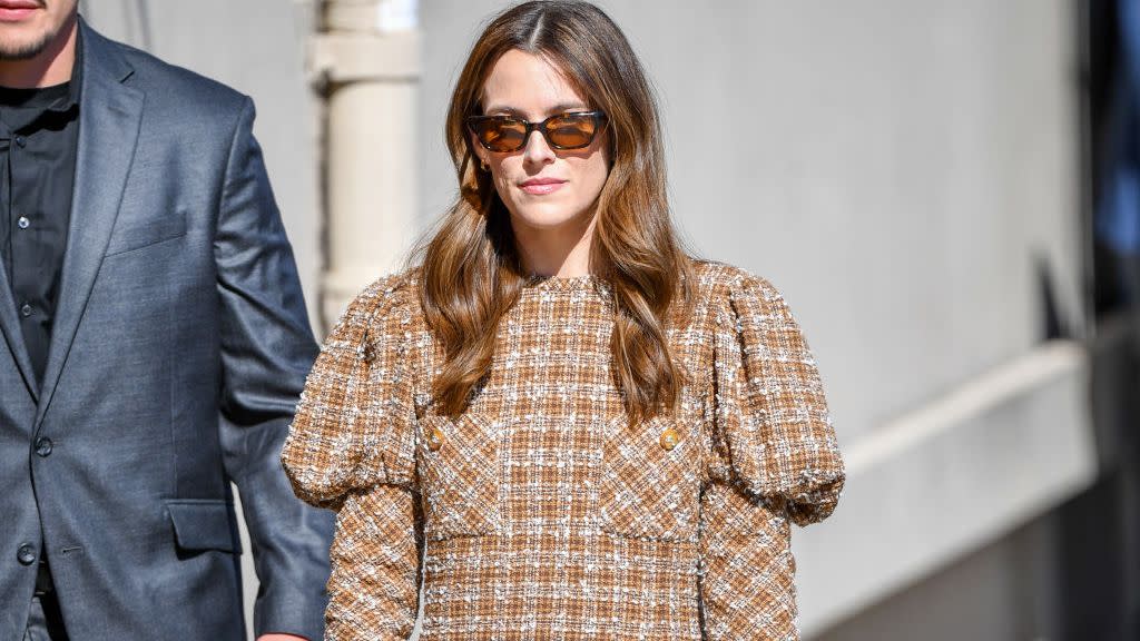 riley keough walks outside, she wears sunglasses and an orange and cream colored plaid dress