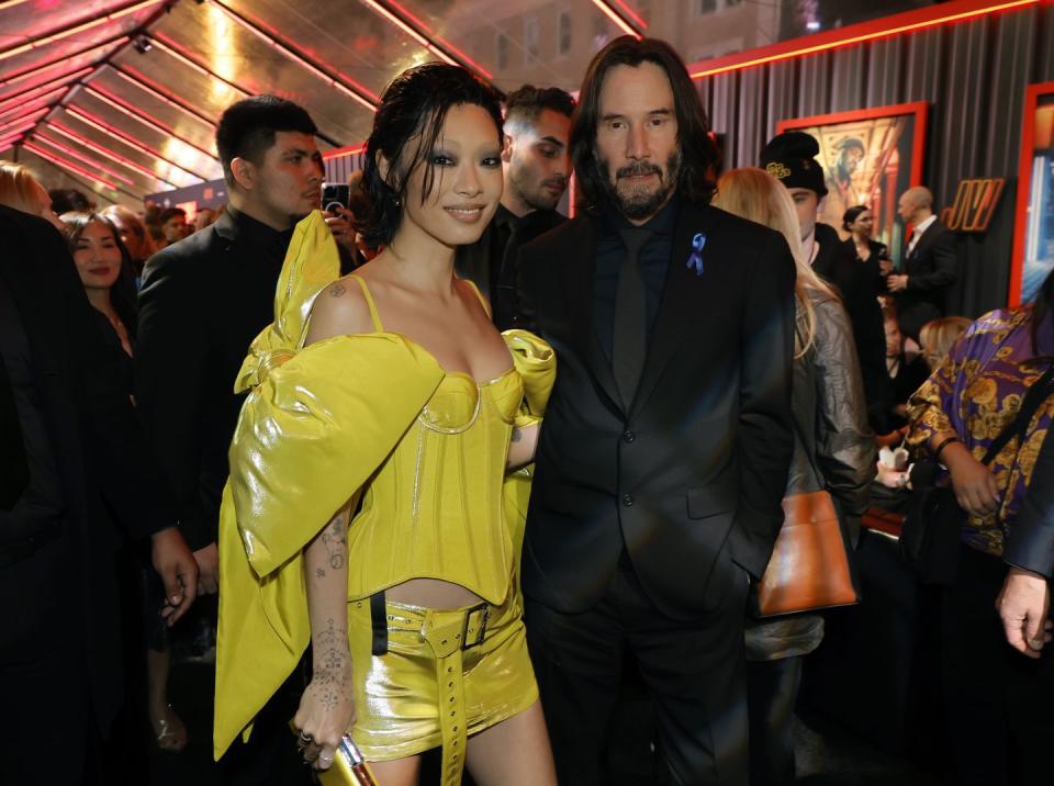 rina sawayama wearing a yellow dress, and keanu reeves wearing a black suit and tie, in a crowded movie theater