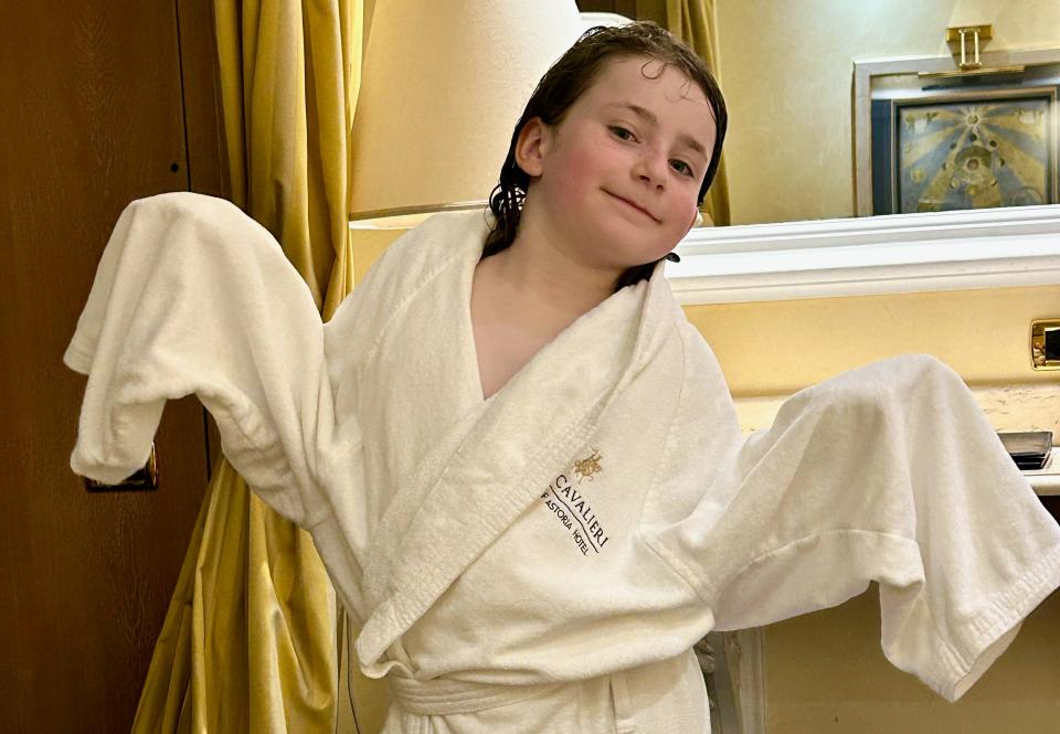 A young girl wearing a large white robe with a Robe Cavalieri logo on it. The sleeves of the robe hang off her and she smiles