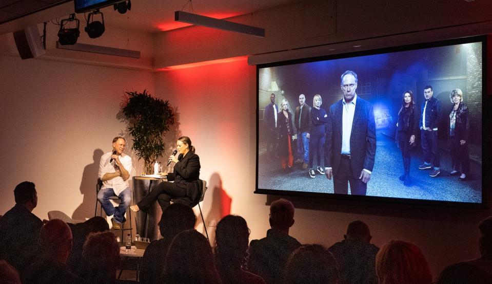 todd boyce sitting at a table with a woman next to a screen showing himself in character as stephen reid on coronation street, displayed in front of a crowd of fans