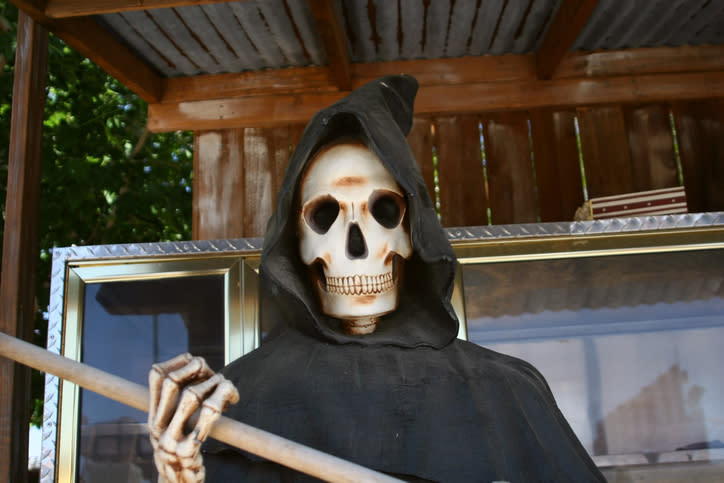 Figure dressed as the Grim Reaper with a skull mask and hooded cloak, near a reflective surface