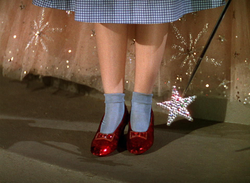 Close-up of Judy Garland's feet from "The Wizard of Oz" wearing blue socks, ruby slippers, standing on a yellow brick road, holding a star-shaped wand