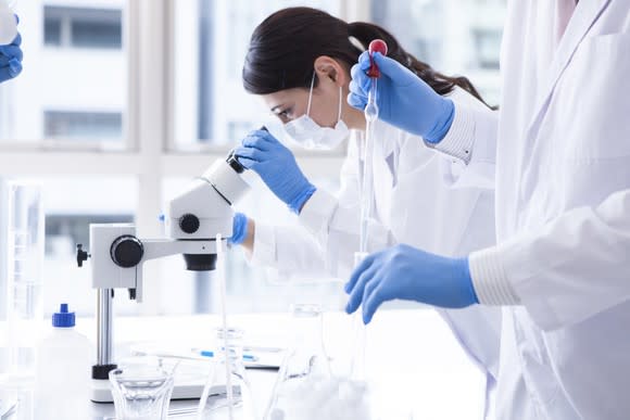 Female scientist in white lab coat looking through microscope with another scientist holding a dropper.
