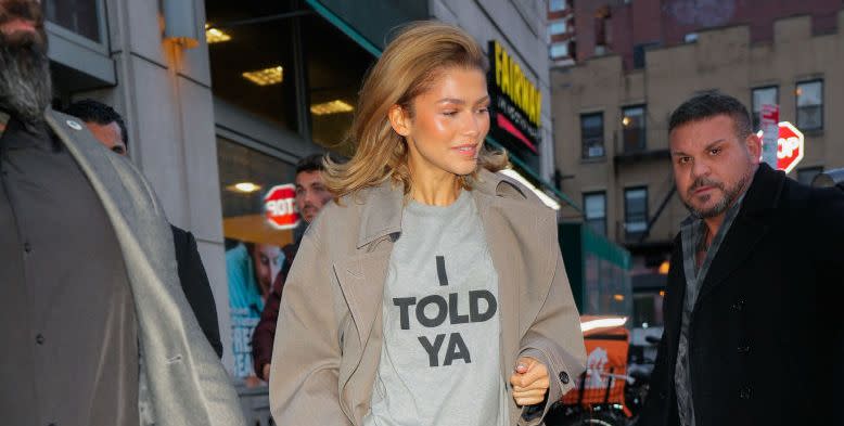 zendaya wearing loewes i told ya t shirt in the colour grey and black lettering with a beige trench coat on top and black trousers and cream shoes