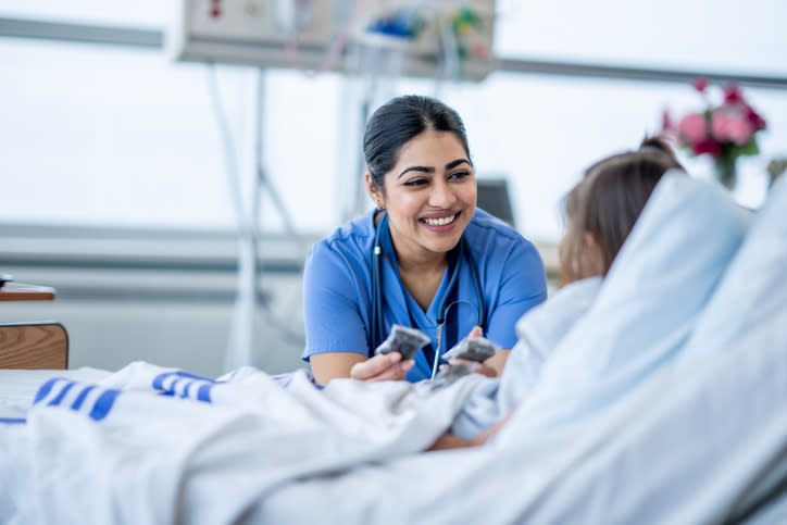 A nurse in blue scrubs is smiling and showing two cards to a patient lying in a hospital bed. The interaction appears positive and caring