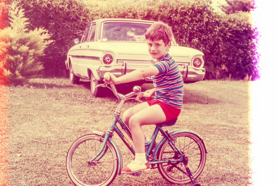 Child in striped shirt and shorts riding a bicycle on a lawn with a vintage car in the background