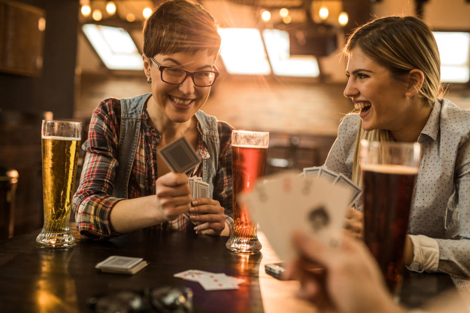 Two women smile and laugh while playing cards and drinking beer in a bar