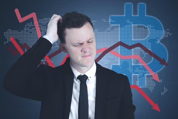 Man scratching his head in front of a large bitcoin logo and several falling arrows.