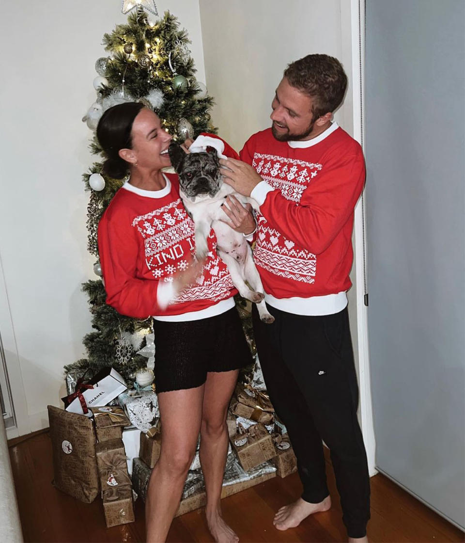 Sam and Bonnie wear matching red Christmas sweaters, are standing in front of a decorated Christmas tree and Bonnie is holding a dog. Sam is trying to put a Santa hat on the dog.