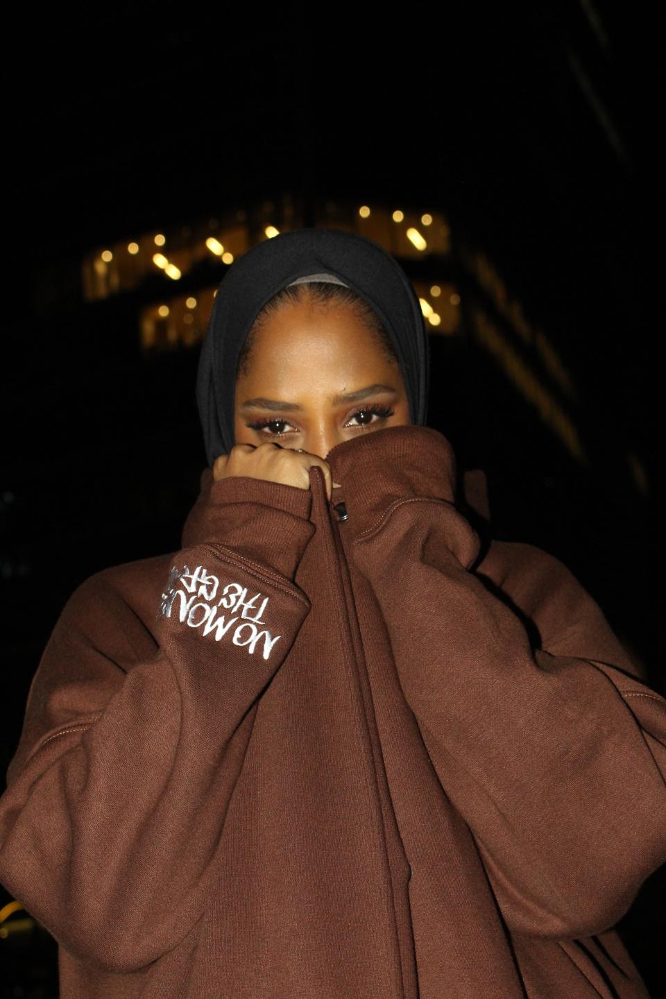 A woman in a hoodie abaya covering lower face with her hands, standing in the night with lights in the background