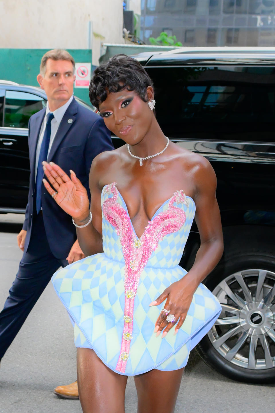 Jodie Turner-Smith is walking, waving, wearing a strapless, structured dress with a checkered pattern. A man in a suit is seen in the background