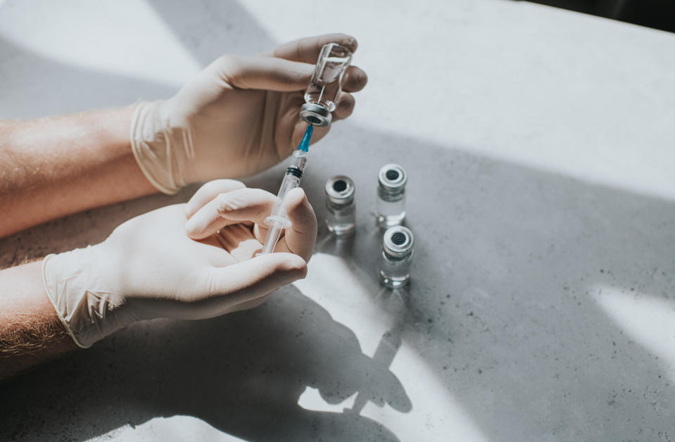 A hand wearing latex gloves hold a phial of clear liquid and a syringe. The medical professional uses the syringe to extract the medicine from the glass bottle. 