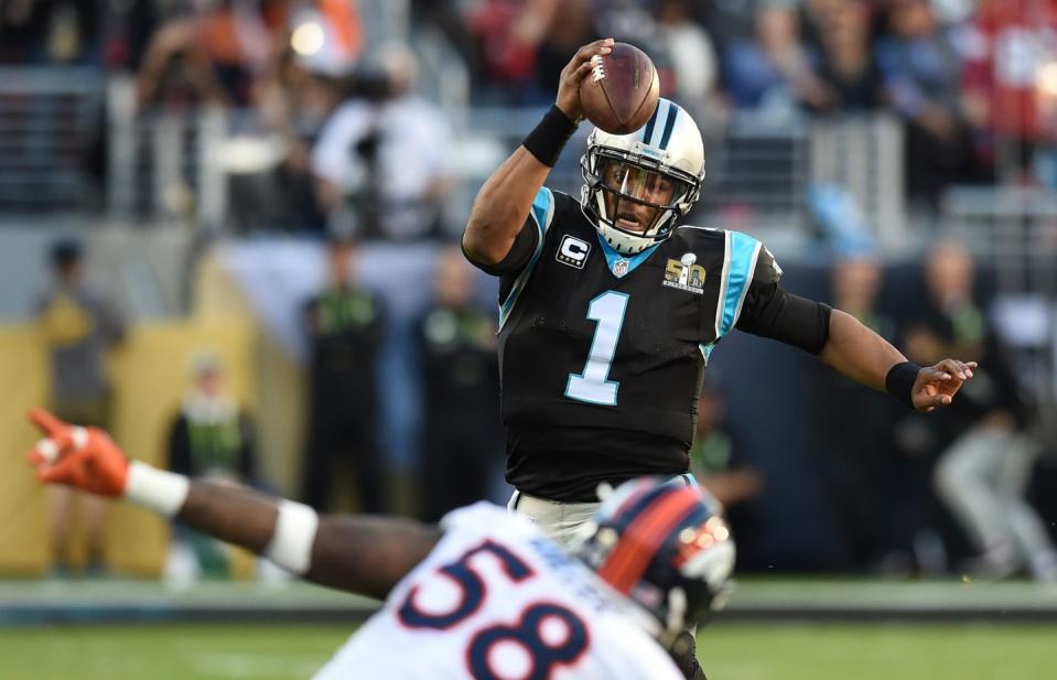 cam newton runs forward with the football while opponent von miller attempts to tackle him, both men wear football uniforms including helmets