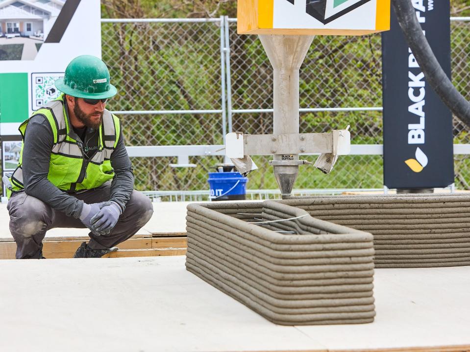 A 3D printer printing concrete onto a wall. A person is kneeling next to the printer.