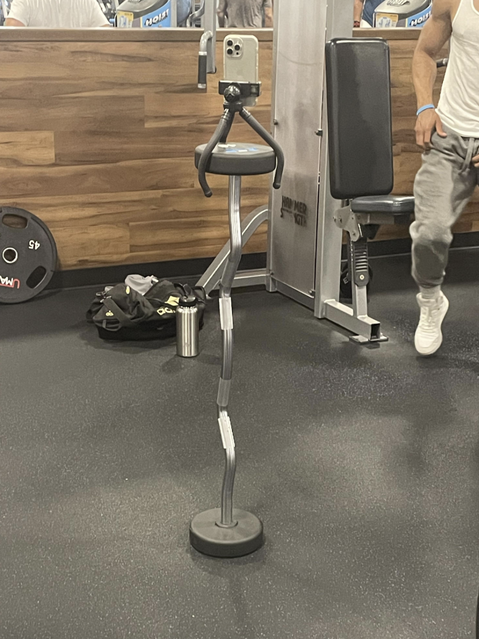 A smartphone mounted on a flexible tripod stand is set up in a gym, with a person partly visible in the background. Gym equipment and a bag are also in sight