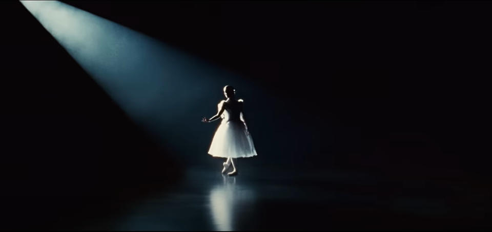 Natalie Portman as Nina in Black Swan, in a spotlight dancing gracefully alone on a dark stage. She wears a knee-length, tulle ballet dress and pointe shoes