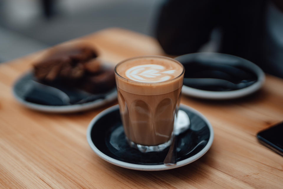 A glass of latte with leaf-shaped foam art on a saucer, accompanied by plates of pastries in the background on a wooden table