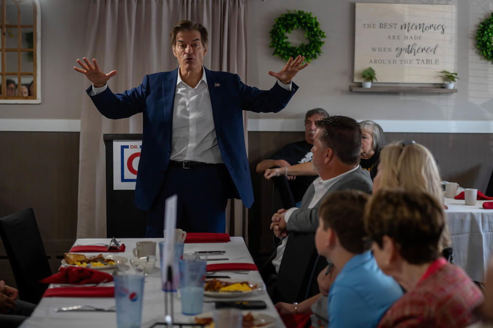 Mehmet Oz stands at a table as people, seated, look on.