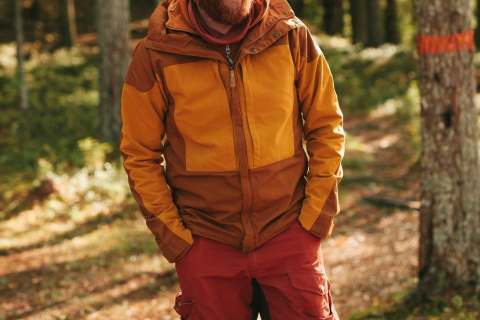A man walks in the forest wearing a lightweight jacket with brown and light orange color pannels.