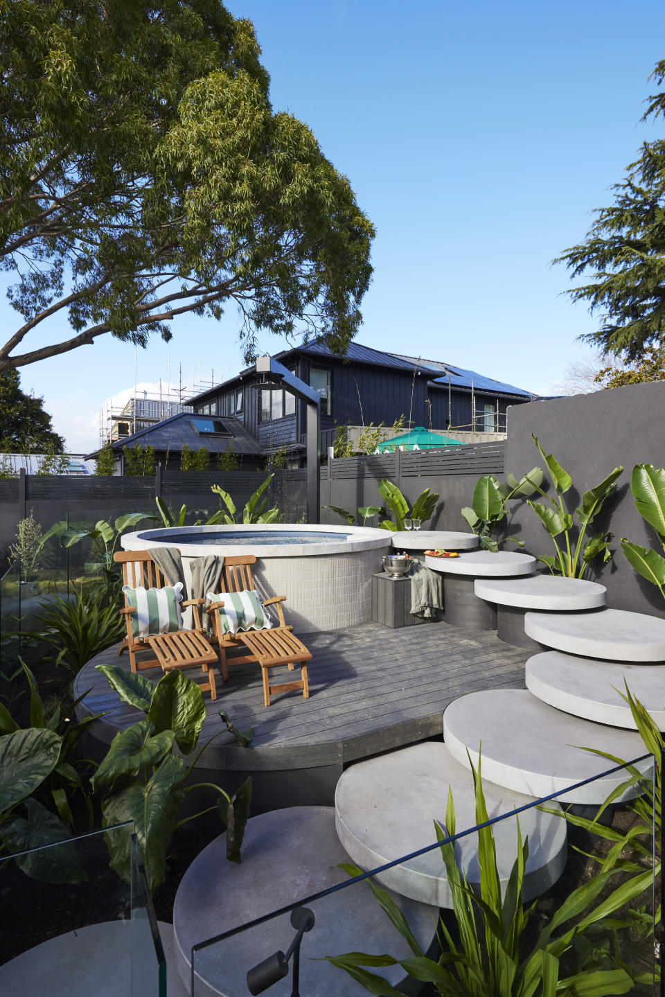 A wide shot of the plunge pool with a series of connected round steppers leading to it. Two wooden pool chairs on the left and plants surrounding.