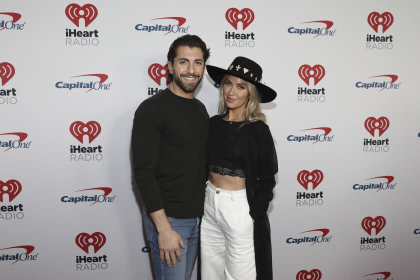 Jason Tartick in a black sweater and jeans next to Kaitlyn Bristowe in a black wide-brimmed hat, sweater and white jeans