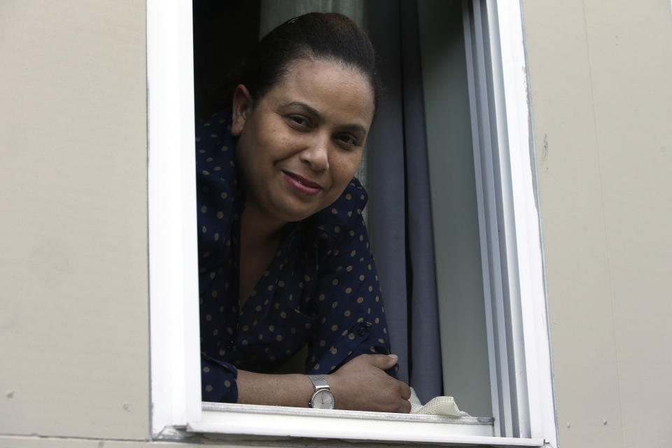 In this Wednesday, May 20, 2020, photo, Wendy De Los Santos, originally from Dominican Republic, stands for a photograph in a window of her home, in Malden, Mass. The 38-year-old Massachusetts resident passed her test to become a U.S. citizen in mid-March, the day before businesses and government offices across her state were ordered shutdown over the spread of the coronavirus. (AP Photo/Steven Senne)