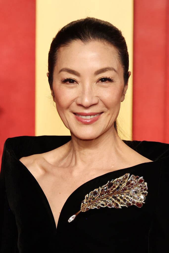 Michelle Yeoh in an off-shoulder black dress, adorned with a jeweled feather brooch, smiling on the red carpet