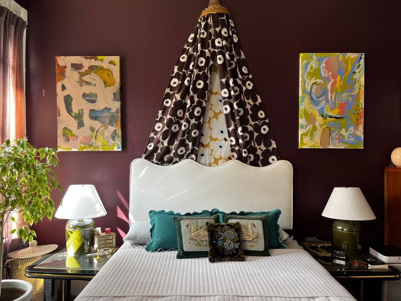 Marimekko fabric draped above white bed with white bedding in eggplant bedroom with colorful artwork.