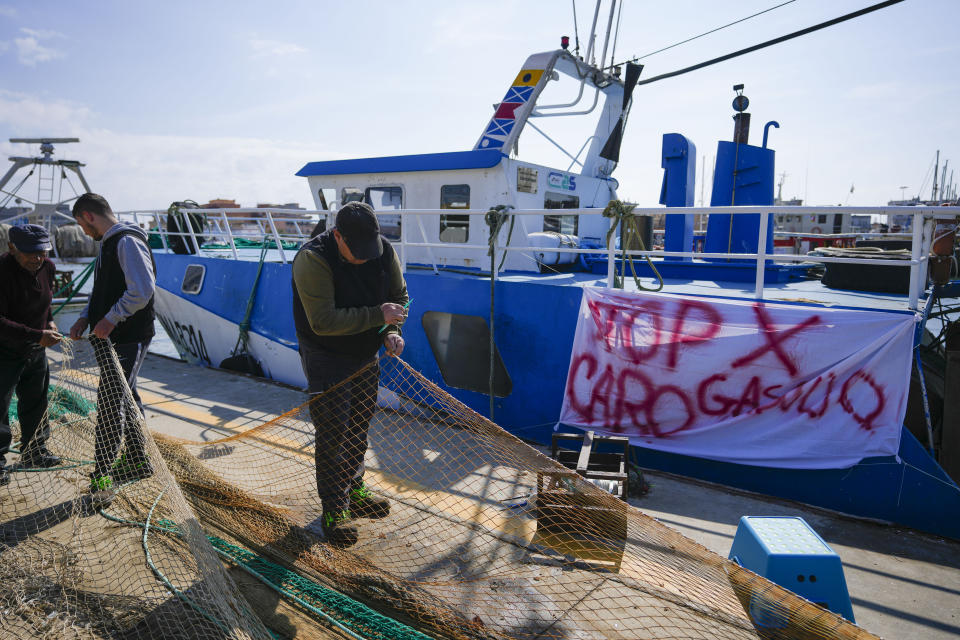Fishermen mend nets in front of his fishing boat with a banner reading "stop for gasoline increase", in the Roman port of Fiumicino, Friday, March 11, 2022. Fishermen, facing huge spikes in oil prices, stayed in port, mending nets instead of casting them. Nowhere more than in Italy, the European Union’s third-largest economy, is dependence on Russian energy taking a higher toll on industry. (AP Photo/Andrew Medichini)