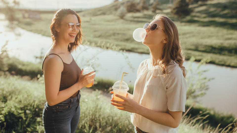 two beautiful girlfriends have fun blowing up a bubble from chewing gum, drinking orange juice in plastic cup, in sunglasses, summer, at sunset, positive facial expression, outdoor.