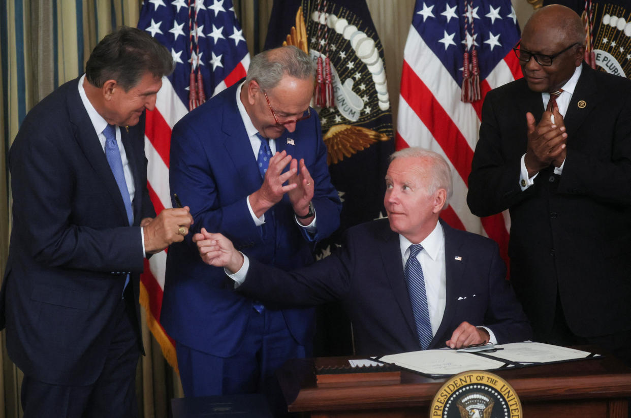 Biden, seated, hands his signing pen to Senator Joe Manchin as Senate Majority Leader Chuck Schumer and House Majority Whip Jim Clyburn look on.