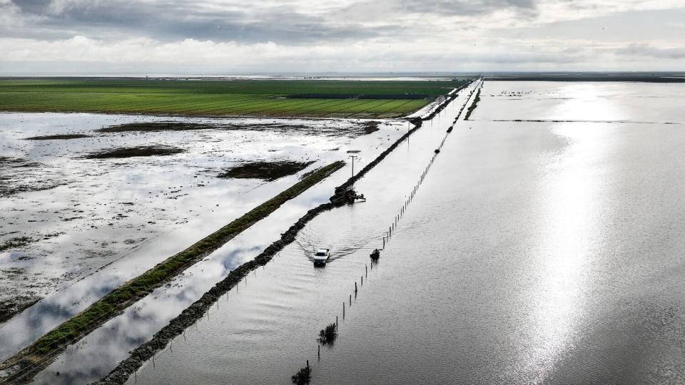 A vehicle drives along a flooded road in an agricultural area.