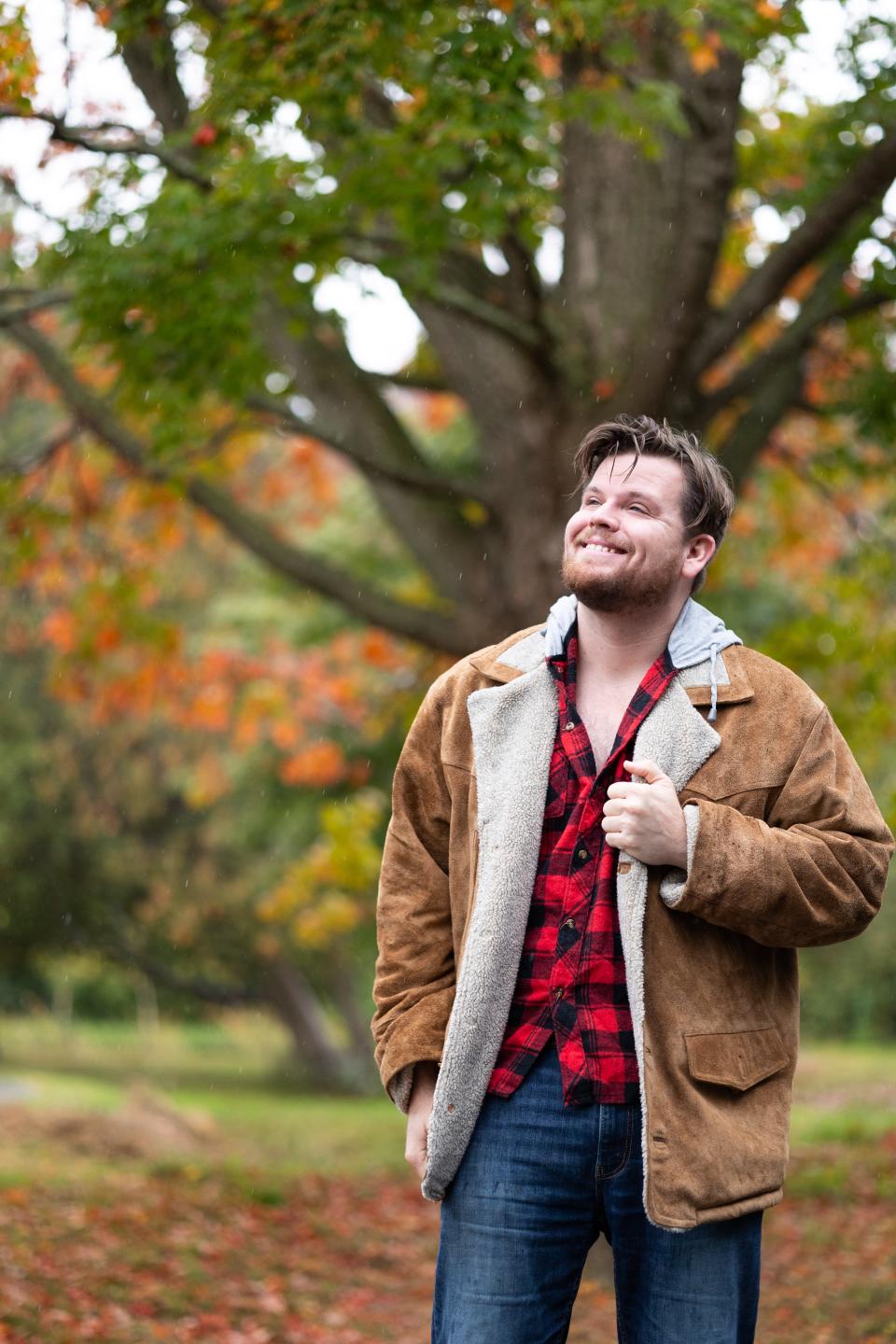 Luke stands in front of a tree, clutching the left lapel of his jacket and smiling.