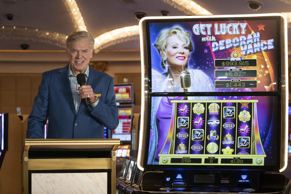 'Hacks' Episode 1, Season 3: a shot of Marty (Christopher Mcdonald) at a lectern with a Deborah Vance themed slot machine to his right. 