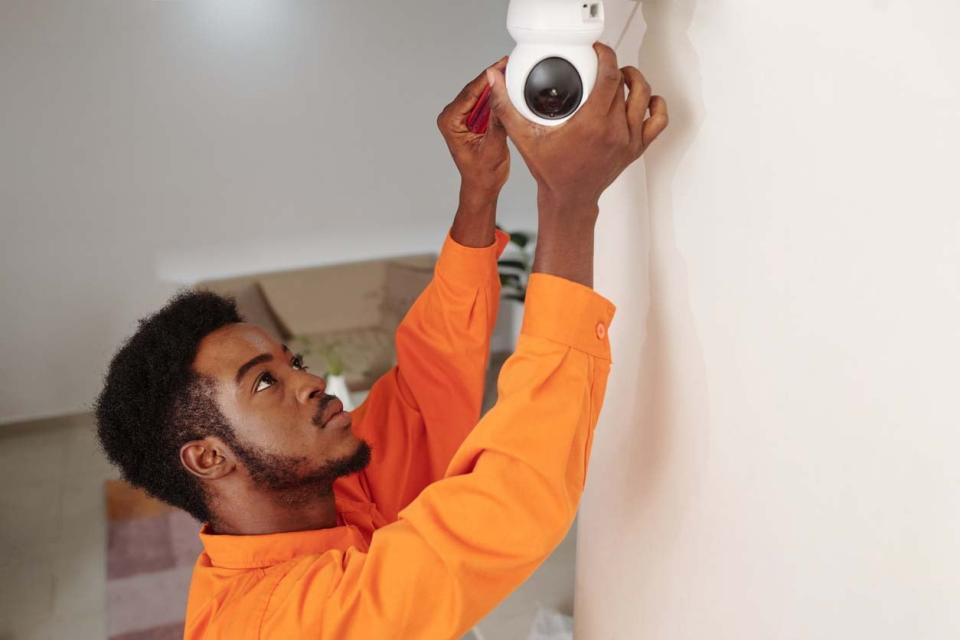 A man in an orange shirt installs a home security camera. 