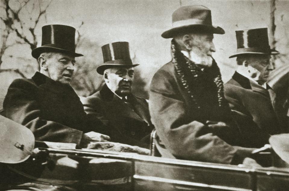 Four middle-aged, white men wearing hats and dark overcoats are taking a ride in a car.