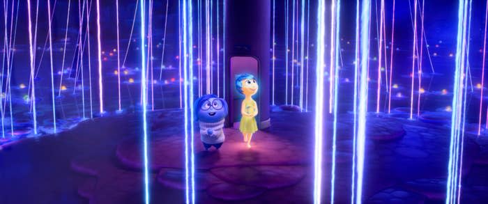 Sadness and Joy from Inside Out 2 stand together in a vibrant, futuristic room illuminated by beams of light