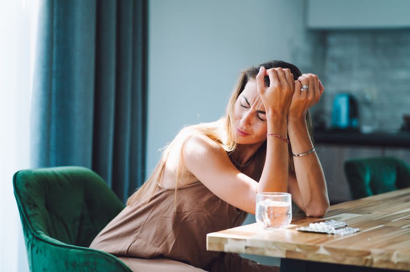 Crying blonde woman with headache  sits hunched over a table, her face filled with sorrow. Water and pills near on the table