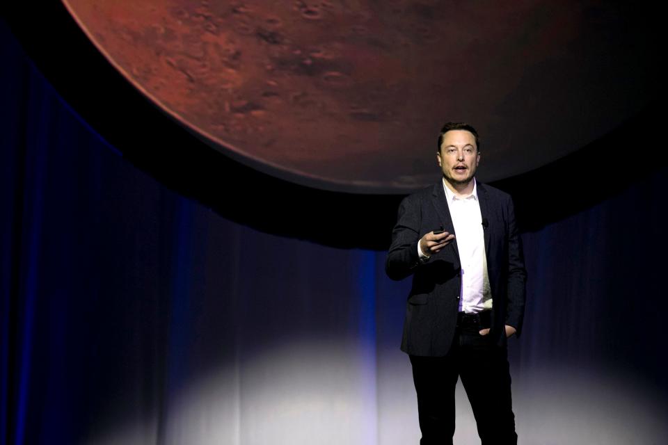 elon musk in suit without tie speaks onstage with an image of mars behind him