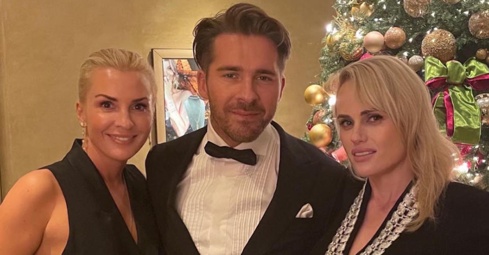 Ramona Agruma, Hugh Sheridan and Rebel Wilson pose for a photo all wearing black in front of a Christmas tree.