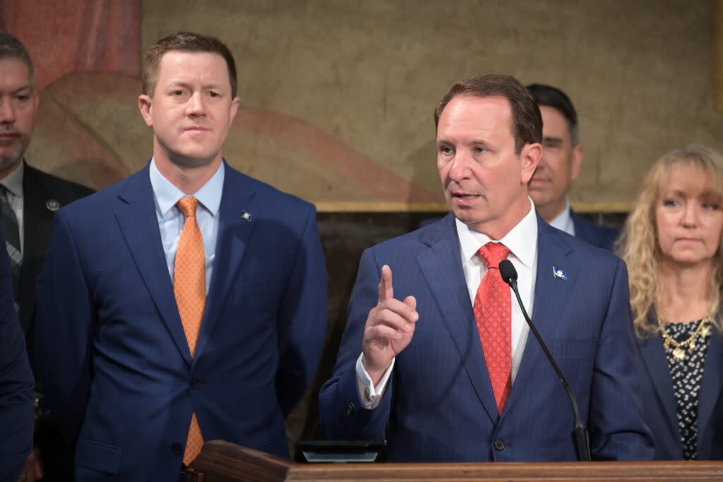 Two men stand side by side. They are both wearing blue suits. One is in front of a microphone and speaking.