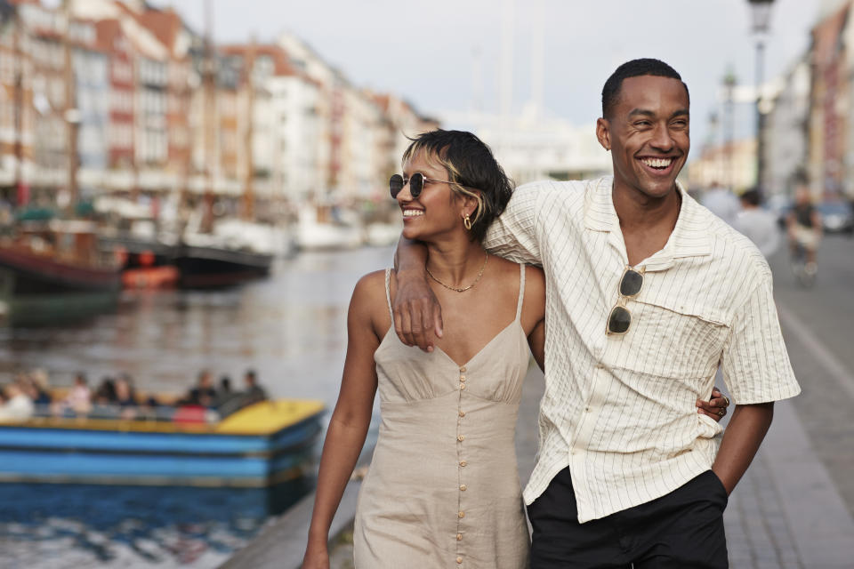 Happy young girlfriend and boyfriend walking with arms around on pier while on vacation, sightseeing