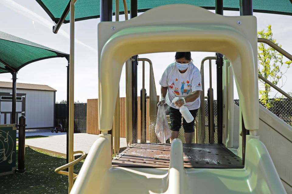 In this May 27, 2020 photo, Ana, a worker at the Frederickson KinderCare daycare center in Tacoma, Wash., wears a mask as she cleans playground equipment following use by a class, a task that is repeated several times a day. In a world weary of the coronavirus, many working parents with young children are now struggling with the decision on when or how they'll be comfortable returning to their child care providers. Frederickson KinderCare has been open throughout the pandemic to care for children of essential workers. (AP Photo/Ted S. Warren)